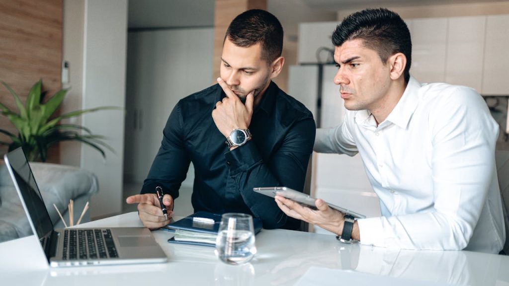 Two men working closely at a laptop, one offering support and guidance, symbolizing self-compassion for men and the care they often extend to others.