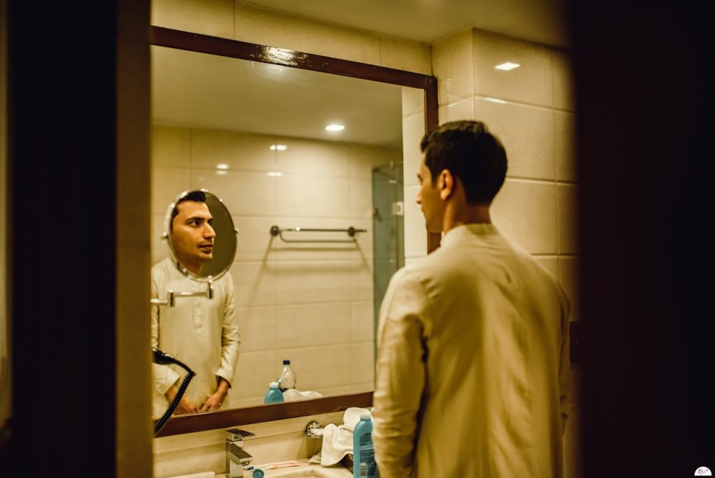 Man looking at his reflection in a bathroom mirror with a thoughtful expression, illustrating men and self-esteem through honest self-reflection.