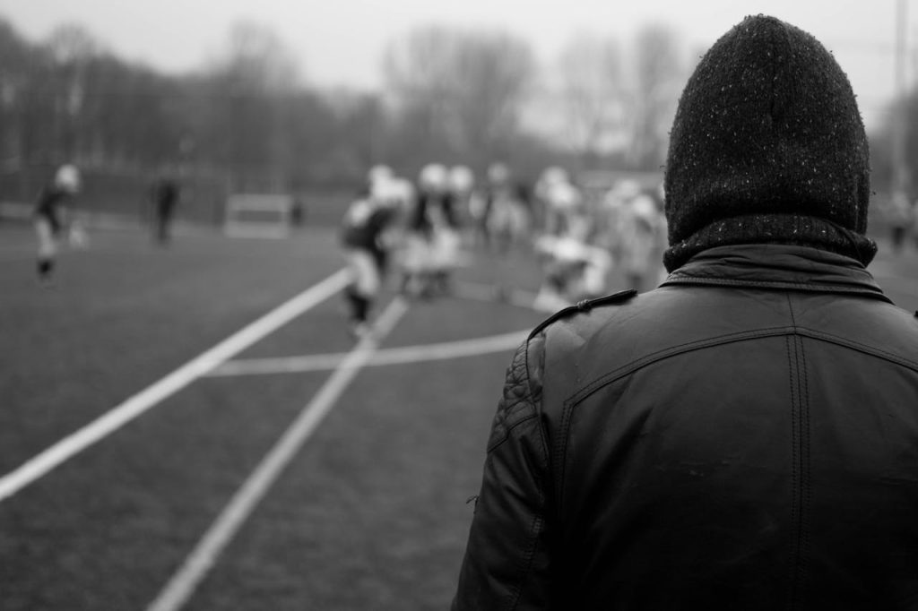Man standing on the sidelines of a sports field watching others play, symbolizing fear of failure in men and self-protective avoidance.