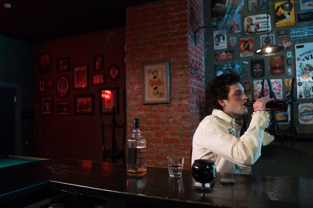A man sitting alone at a dimly lit bar, drinking from a bottle and looking distant, symbolizing unhealthy coping and avoidance instead of finding inner peace.