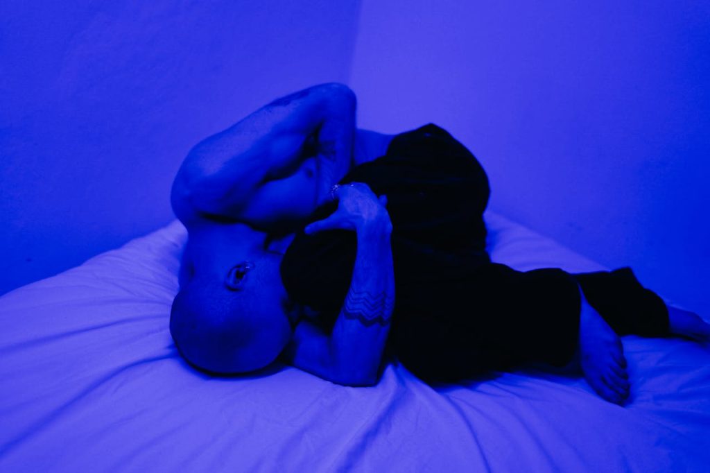 A man lying curled on a bed under blue lighting, conveying isolation, emotional withdrawal, and psychological distress—common signs of depression in men.