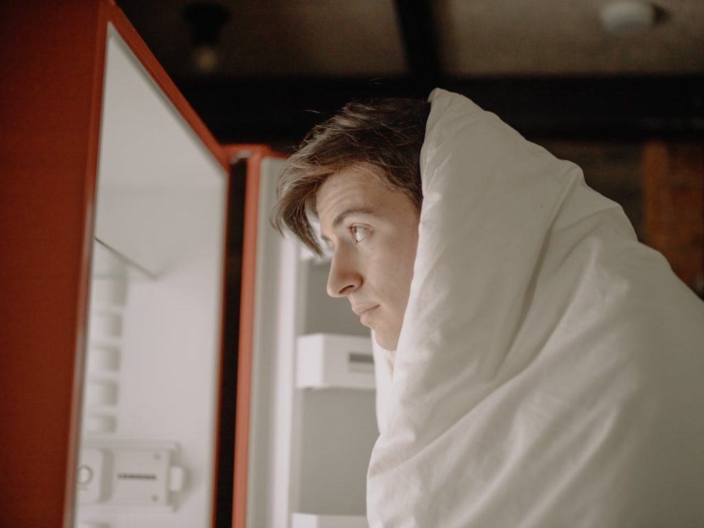 A man wrapped in a blanket standing in front of an open refrigerator at night, appearing absent-minded and fatigued, illustrating disrupted appetite and sleep patterns associated with depression in men.