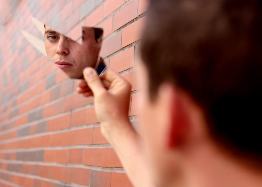 A man looking at his reflection in a broken mirror, appearing emotionally distant, illustrating self-recognition and inner emptiness as signs of depression in men.
