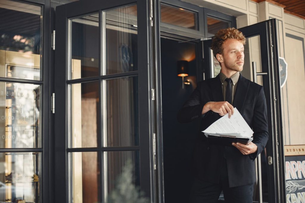 Man in a suit standing at the entrance of a formal building holding documents, symbolizing imposter syndrome in men and the question of belonging in high-status environments.