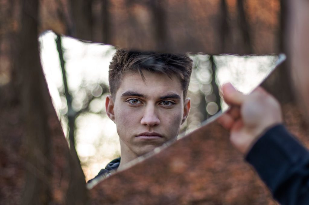 A young man looking at his reflection in a broken mirror, appearing serious and guarded, symbolizing how unresolved childhood trauma in men shapes present identity.