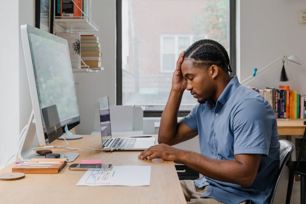[Image Placement 1: "A man sitting at a desk with his head in his hands, surrounded by work - not dramatic despair, but the quiet, grinding exhaustion of someone who's been their own harshest critic for too long."]