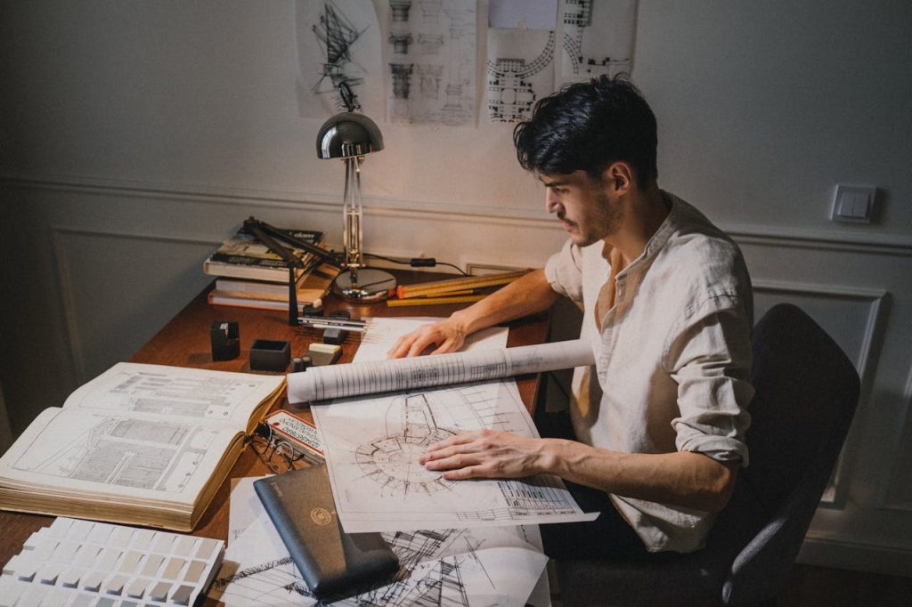 Man working late at a desk surrounded by drafts and architectural plans, symbolizing fear of failure in men and perfectionistic over-refinement.