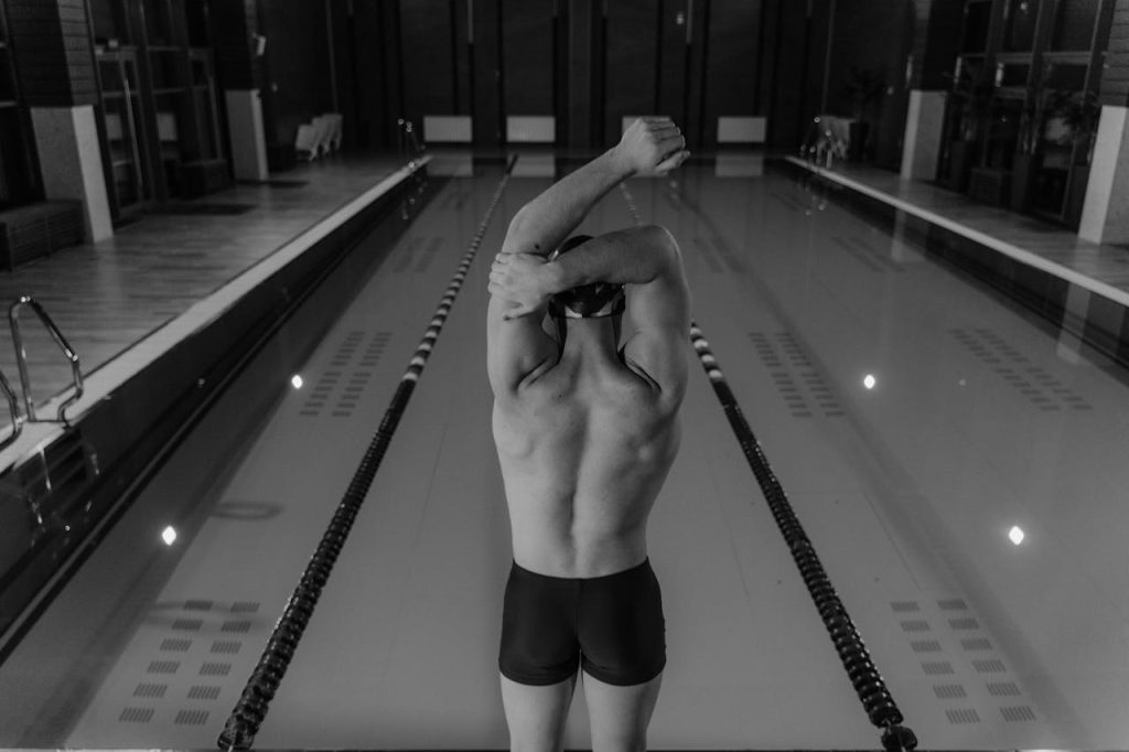Man standing at the edge of an indoor swimming pool lane, stretching before a dive, symbolizing fear of failure in men and hesitation at the point of decision.