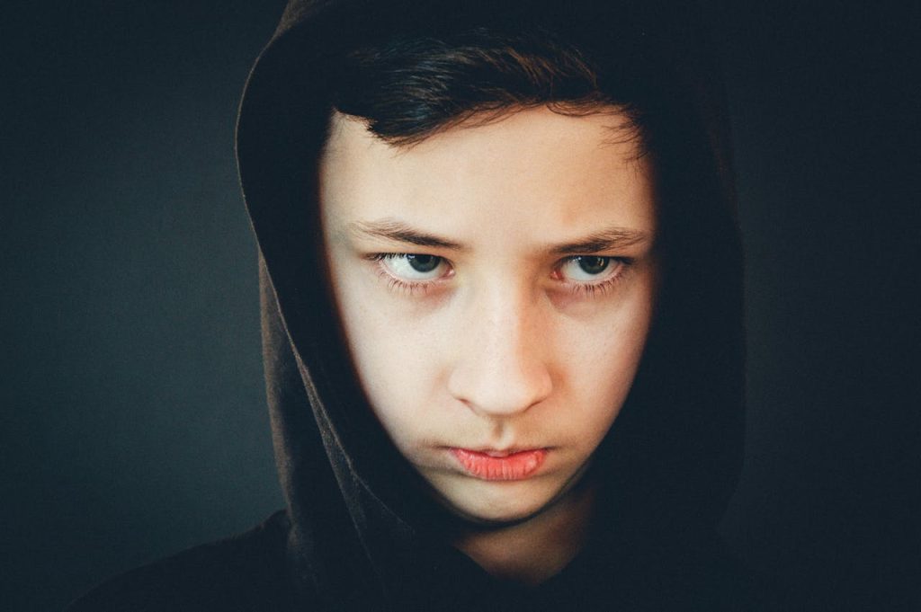 A teenage boy wearing a dark hood, staring forward with a tense and hollow expression, illustrating the psychological impact of unresolved childhood trauma in men.