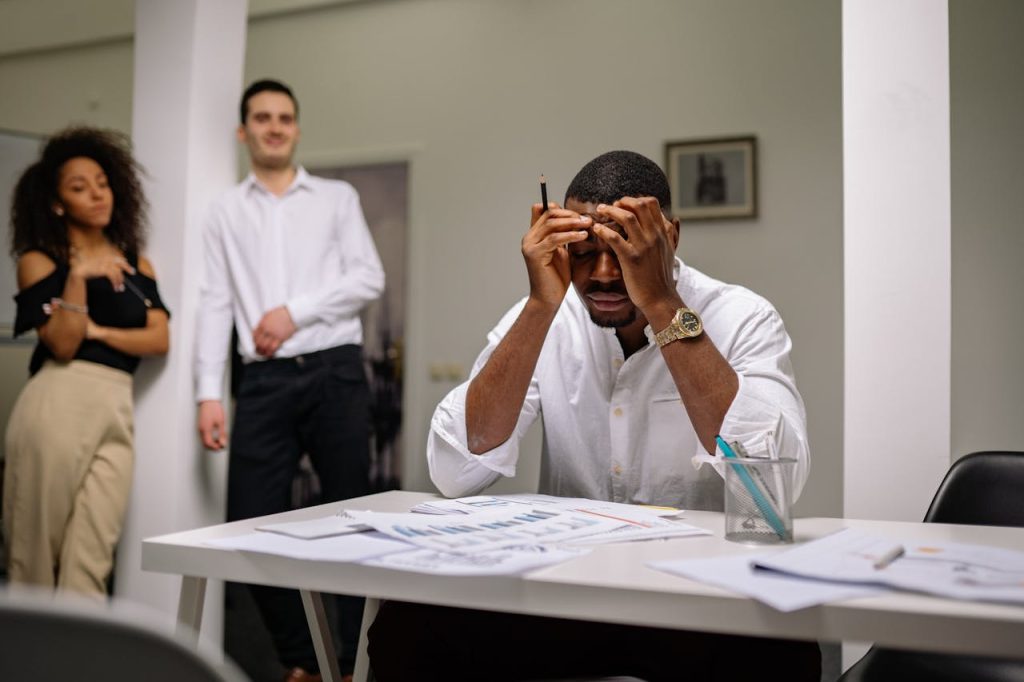 Man sitting at a desk holding his head in visible stress while colleagues stand behind him, illustrating the fragile gap between performance-based confidence and men and self-esteem.